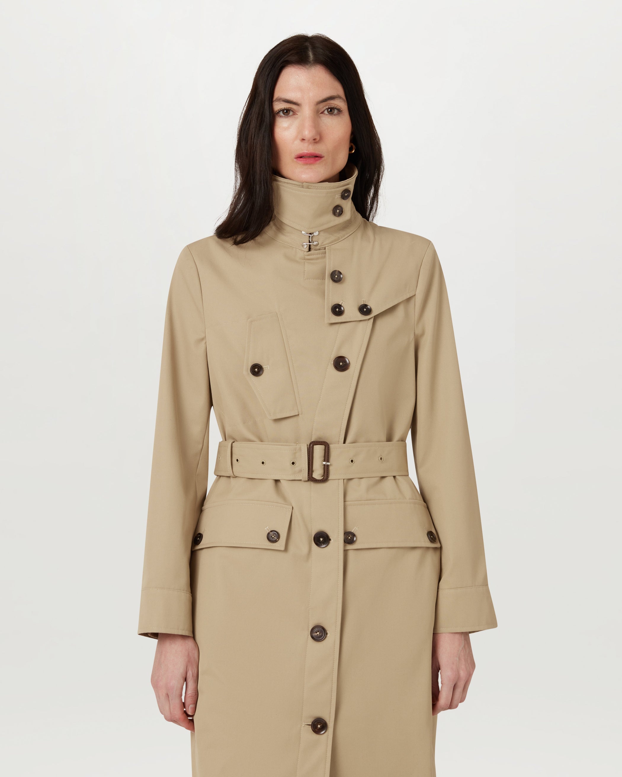Model wearing TT Trench Coat in Light Sand with high collar reverse front opening and belted waist.